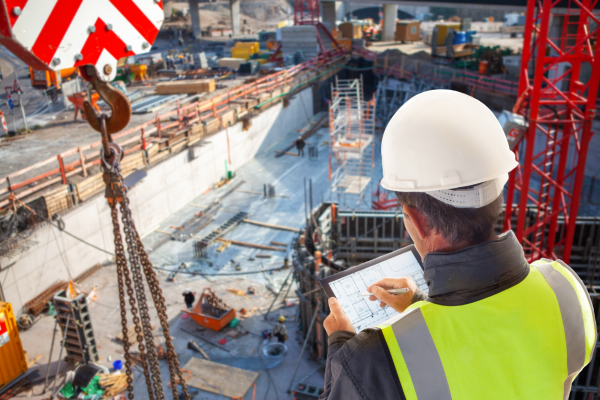 A construction worker in a hardhat and safety vest reviews plans at a building site with cranes, scaffolding, and various construction materials in the background.