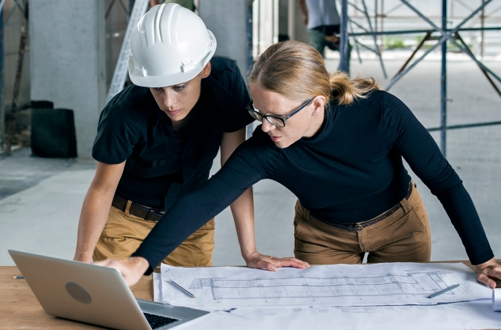 Two people reviewing blueprints on a table; one wearing a hard hat, the other pointing at a laptop screen. Both are in a construction site.