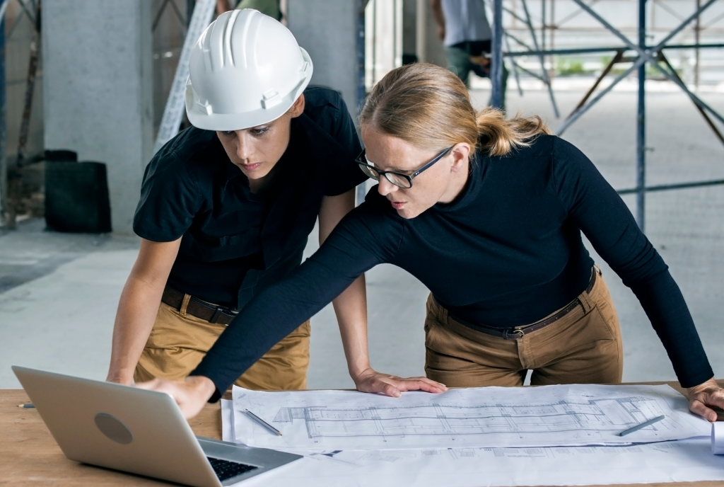 Two people reviewing blueprints on a table; one wearing a hard hat, the other pointing at a laptop screen. Both are in a construction site.