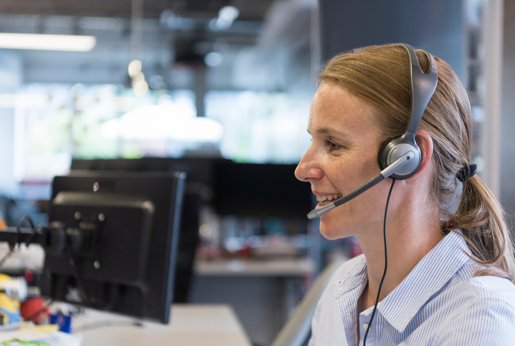 A woman with a headset is smiling and looking at a computer screen in a modern office environment.