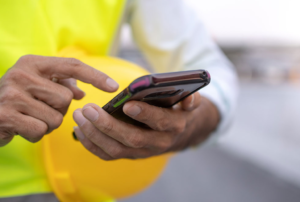 A person in a bright yellow safety vest uses their smartphone while holding a yellow hard hat.