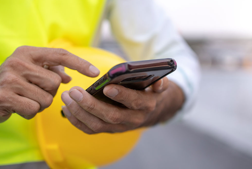 A person in a bright yellow safety vest uses their smartphone while holding a yellow hard hat. The focus is on the hands and the phone.