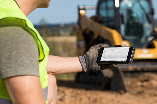 A construction worker wearing a reflective vest and gloves views information on a smartphone at a construction site with a blurred yellow excavator in the background.