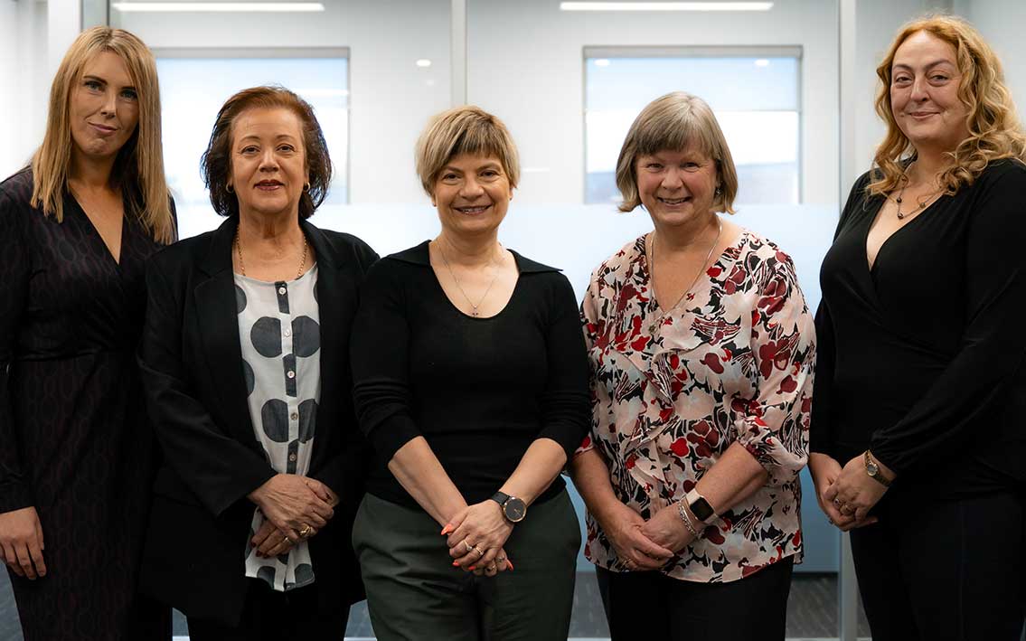 Five women standing indoors, all facing the camera and smiling.