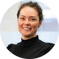 Person with curly hair smiles while wearing a black turtleneck. They are in a well-lit room with a soft-focus background.