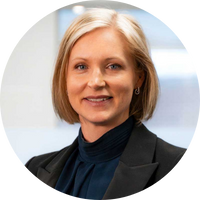 A person with short blonde hair wearing a dark blazer and blouse, standing indoors with a blurred background.