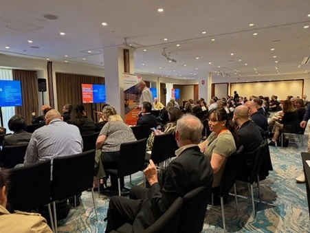 Attendees sit in rows facing screens and a speaker at a conference or seminar held in a well-lit room. Attendees sit in rows facing screens and a speaker at a conference or seminar held in a well-lit room.